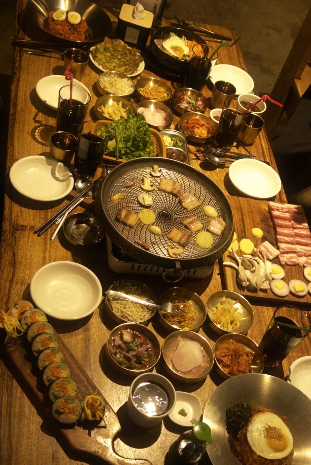An overhead view of a full Korean BBQ spread on a wooden table featuring a round charcoal grill with meat and mushrooms cooking, raw pork belly slices, multiple banchan side dishes, kimbap rolls, bibimbap and dark drinks.